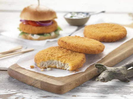 Traditional breaded fishcake on a wooden chopping board with a fishburger in the background.